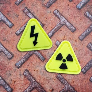 Op de afbeelding: Twee driehoekige patches met een heldere neongele achtergrond en verhoogde zwarte symbolen. Eén patch heeft een zwart bliksemschichtpictogram, terwijl de andere patch een zwart stralingsgevaarsymbool toont. Beide items hebben een gladde afwerking.