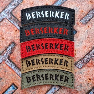 Puede incluir: Cinco parches de tela negra, roja, marrón y verde con la palabra "BERSERKER" en letras blancas, rojas, negras y negras, respectivamente. Los parches son curvos y tienen un borde cosido.