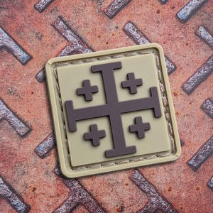 May include: A square patch featuring a Jerusalem cross design. The cross is dark gray, set against a tan background, and framed by a light tan border. The patch is on a textured, rusty metal surface.
