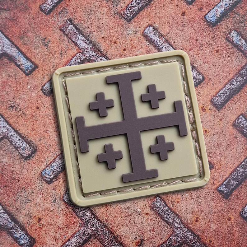 May include: A square patch featuring a Jerusalem cross design. The cross is dark gray, set against a tan background, and framed by a light tan border. The patch is on a textured, rusty metal surface.