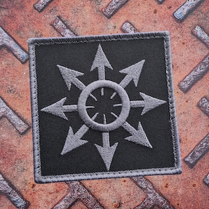 Könnte beinhalten: Quadratischer schwarzer Aufnäher mit einem grau gestickten Symbol, das einen zentralen Kreis mit strahlenförmigen Pfeilen zeigt. Der Aufnäher hat einen grauen Rand und ist vor einem rostigen Metallhintergrund platziert.