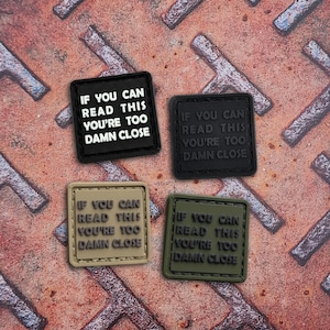 May include: Four square patches with the text "IF YOU CAN READ THIS YOU'RE TOO DAMN CLOSE". The patches are black, dark grey, tan, and olive green. The patches are on a textured, rusty metal surface.