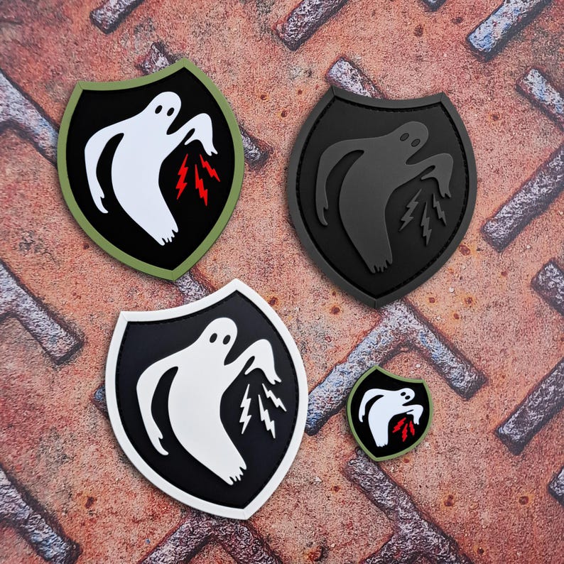 Op de afbeelding: Vier schildvormige patches met een spookontwerp. De patches zijn zwart, wit en olijfgroen, met een witte geest en rode bliksemschichten. De patches vari&euml;ren in grootte en worden weergegeven op een roestig metalen oppervlak.