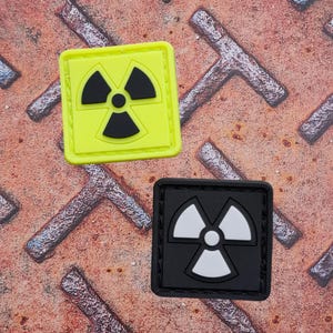 以下が含まれることがあります： 放射能マークの2つの正方形パッチ。1つは黒いシンボルがネオンイエローの背景に、もう1つは白いシンボルが黒い背景にあります。それらは、質感のある錆びた金属の表面に配置されています。