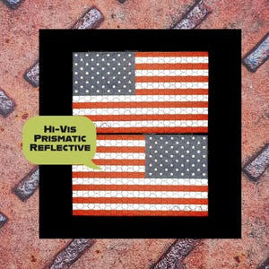K&ouml;nnte beinhalten: Zwei reflektierende amerikanische Flaggen-Patches mit roten und wei&szlig;en Streifen und einem Feld wei&szlig;er Sterne auf grauem Hintergrund. Die Patches sind schwarz umrandet, mit dem Text "Hi-Vis Prismatic Reflective" in einer gr&uuml;nen Sprechblase.