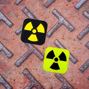 Può includere: Due toppe quadrate con un simbolo di radiazione. Una è nera con un simbolo giallo, l'altra è verde neon con un simbolo nero. Sono su una superficie metallica arrugginita.
