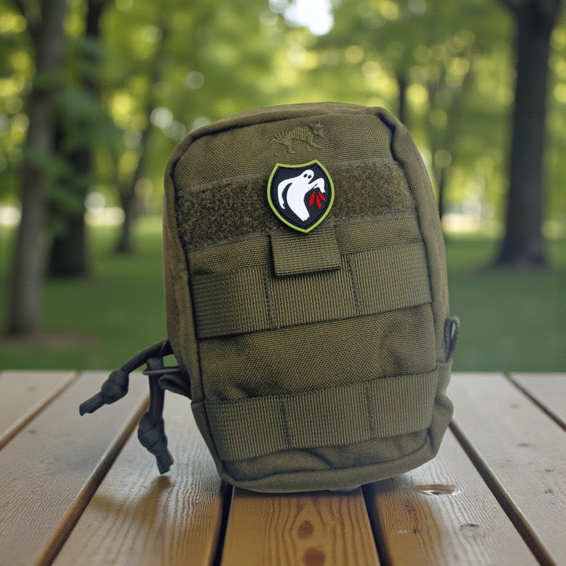 Op de afbeelding: Olijfgroene tactische tas met een zwart-witte spookpatch met een rode vlam. De tas heeft meerdere singelbanden en een klittenbandsluiting. Het ligt op een houten oppervlak.