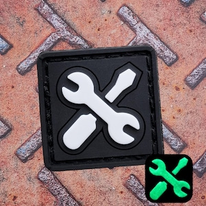 以下が含まれることがあります： 白のレンチとドライバーが交差したデザインの黒い正方形のパッチ。パッチは黒い縁取りがあり、質感のある錆びた金属の背景に配置されています。デザインの小さく光る緑色のバージョンが隅にあります。