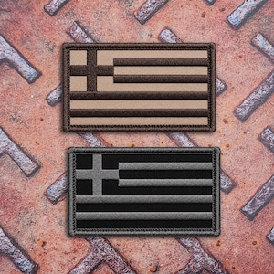 Puede incluir: Dos parches rectangulares con el diseño de la bandera griega. El parche superior presenta un esquema de color beige y marrón oscuro, mientras que el inferior muestra un esquema de color negro y gris. Ambos parches tienen un borde cosido.
