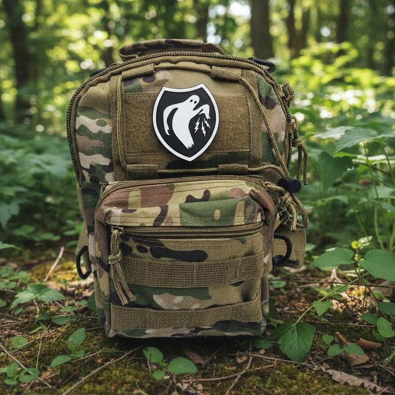 Op de afbeelding: Een camouflage tactische rugzak met meerdere compartimenten en riemen. Een zwart-witte patch met een spook en bliksemschicht is aan de voorkant bevestigd. De tas is ontworpen voor gebruik buitenshuis en wordt getoond in een bosrijke omgeving.