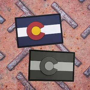 Op de afbeelding: Twee rechthoekige patches met het ontwerp van de vlag van de staat Colorado. Eén patch heeft een marineblauwe achtergrond met een witte streep en een rood en geel 'C'-embleem. De andere patch heeft een grijze achtergrond met een witte streep en een grijs 'C'-embleem.