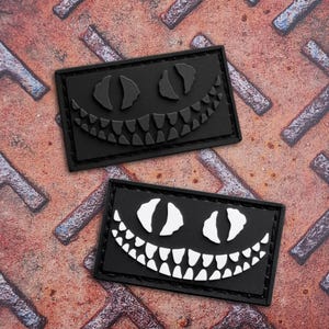 Op de afbeelding: Twee zwarte rechthoekige patches met een gestileerde Cheshire Cat-grijns. Eén patch heeft een zwarte grijns, de andere een witte grijns. De patches liggen op een getextureerde, roestige metalen achtergrond.