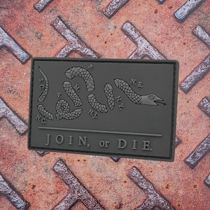 May include: A rectangular, dark gray patch featuring a segmented snake design with the text "JOIN, or DIE." The snake is divided into segments, each labeled with state abbreviations. The patch is set against a textured, rusty metal surface.
