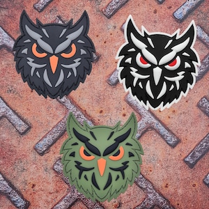 Könnte beinhalten: Drei Eulen-Patches in verschiedenen Farbschemata. Die erste Eule ist schwarz mit grauen Details und orangefarbenen Augen. Die zweite Eule ist schwarz mit weißen Details und roten Augen. Die dritte Eule ist grün mit schwarzen Details und orangefarbenen Augen.