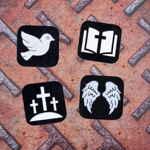 Può includere: Quattro toppe quadrate nere con simboli religiosi bianchi. Una toppa presenta una colomba, un'altra un libro aperto, una terza ha tre croci e l'ultima mostra un paio di ali. Le toppe sono su una superficie metallica arrugginita e strutturata.