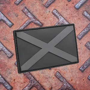 Könnte beinhalten: Ein rechteckiger Aufnäher mit einem grauen X-Design auf schwarzem Hintergrund. Der Aufnäher hat einen schwarzen Rand und befindet sich auf einer strukturierten, rostigen Metalloberfläche. Das Design ist einfach und grafisch.