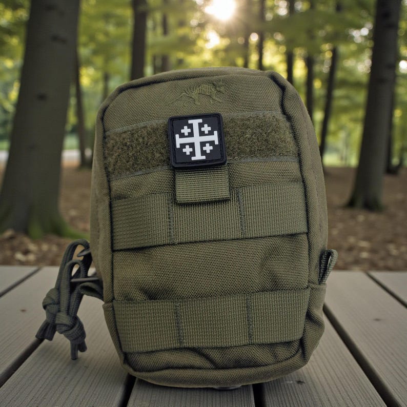 May include: Olive green tactical pouch with a black and white patch featuring a Jerusalem cross. The pouch has multiple straps and a loop for attachment. The background is a blurred forest scene.