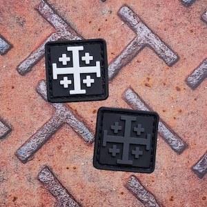 Op de afbeelding: Twee vierkante zwarte patches met het Jeruzalemkruisontwerp. Eén patch heeft een wit kruis, de andere een grijs kruis. De patches liggen op een getextureerd, roestig metalen oppervlak.