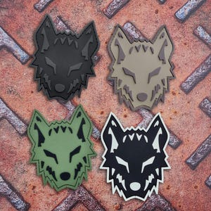 May include: Four wolf head patches in black, tan, green, and black and white. Each patch features a detailed wolf face design with sharp angles and a textured appearance. The patches are arranged on a rusty metal surface.