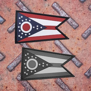 May include: Two Ohio state flag patches. One patch features the flag's red, white, and blue design with a black border. The second patch is a grayscale version of the same flag with a black border. Both patches are rectangular with a unique cut-out design.