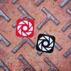 Könnte beinhalten: Zwei quadratische Aufnäher, einer rot und einer schwarz, jeweils mit einem weißen Blenden-Design. Sie liegen auf einer strukturierten, rostigen Metalloberfläche mit einem sich wiederholenden T-förmigen Muster.