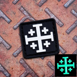 May include: A black square patch featuring a white Jerusalem cross design. The patch has a hook and loop backing. A smaller, glowing green version of the cross is shown in the lower right corner. The patch is on a rusty metal surface.