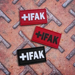 Könnte beinhalten: Drei rechteckige Aufnäher mit dem Text "+IFAK". Zwei Aufnäher sind rot mit weißem und schwarzem Text, einer ist schwarz mit weißem Text. Die Aufnäher liegen auf einer strukturierten, rostigen Metalloberfläche.