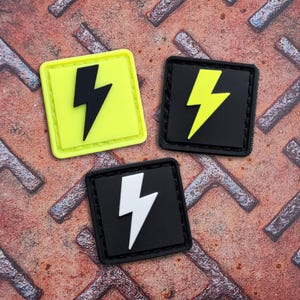 Op de afbeelding: Drie vierkante patches met een bliksemontwerp. Eén patch is neongeel met een zwarte bliksem, een andere is zwart met een gele bliksem en de derde is zwart met een witte bliksem.