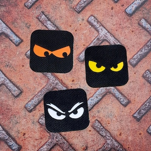 May include: Three black square patches featuring various eye designs. One has orange eyes, another has yellow eyes, and the third has white eyes. The patches are on a textured, rusty metal surface.