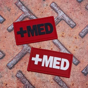 Może przedstawiać: Dwie czerwone prostokątne naszywki z napisem '+MED'. Jedna naszywka ma czarny tekst, druga biały. Naszywki znajdują się na teksturowanej, zardzewiałej metalowej powierzchni.