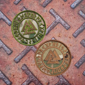 May include: Two circular embroidered patches featuring a camouflage pattern and a central triangular symbol. The patches are bordered by a ring of runes in green and brown thread, set against a textured metal background.