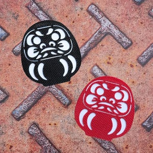 以下が含まれることがあります： 白の顔の特徴と曲がったアクセントを持つ、黒と赤の2つのダルマ人形。人形は、質感のある錆びた金属の背景に配置され、印象的なコントラストを生み出しています。