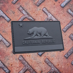 Puede incluir: Un parche rectangular gris oscuro con el diseño de la bandera del estado de California. El diseño incluye un oso, una estrella y las palabras "CALIFORNIA REPUBLIC". El parche probablemente está hecho de un material duradero.