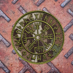 May include: Round patch with a camouflage pattern and green border. The patch features a central symbol with runic inscriptions around the edge. The design is embroidered with green thread.