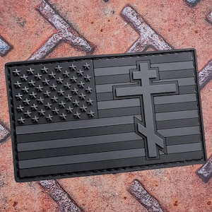 Op de afbeelding: Een zwart en grijs embleem met het ontwerp van de Amerikaanse vlag en een kruis. Het embleem heeft een rechthoekige vorm en een getextureerd oppervlak. De sterren en strepen van de vlag zijn duidelijk zichtbaar, evenals het kruis.