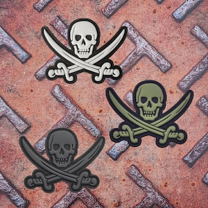 May include: Three pirate skull and crossbones patches in white, olive green, and black. Each patch features a skull over two crossed sabers. The patches are on a rusty, textured metal surface.