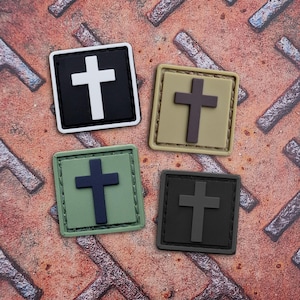 May include: Four square patches with a cross design. The patches are in black, tan, green, and dark gray. Each patch has a different color cross, including white, brown, navy, and gray. The patches are on a textured, rusty metal surface.