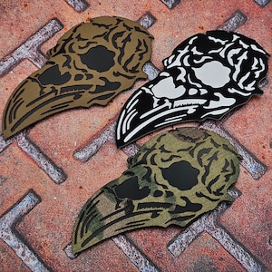 May include: Three patches in the shape of a bird skull. One is tan and black, another is black and white, and the third is camouflage green and black. The patches are on a rusty, textured metal surface.