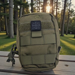 May include: Olive green tactical pouch with multiple straps and a black patch featuring a cross design. The pouch is made of durable material and has a loop for attaching to other gear. The background shows a park with trees and sunlight.