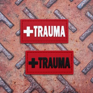 Op de afbeelding: Twee rechthoekige patches met een rode achtergrond en gestikte randen. De bovenste patch heeft een wit medisch kruis en het woord TRAUMA in het wit. De onderste patch heeft een zwart medisch kruis en het woord TRAUMA in het zwart.
