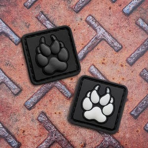 Könnte beinhalten: Zwei quadratische, schwarze Aufnäher mit einem Pfotenabdruck-Design. Ein Aufnäher hat einen schwarzen Pfotenabdruck, der andere einen weißen. Die Aufnäher befinden sich auf einem rostigen, strukturierten Metallhintergrund.