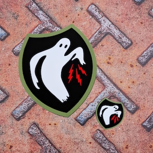 Op de afbeelding: Twee zwarte en groene schildvormige patches. Elke patch heeft een wit spookontwerp met rode bliksemschichten op een zwarte achtergrond. De patches liggen op een getextureerd, roestig metalen oppervlak.