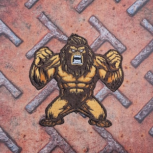 May include: Embroidered patch of a cartoon Bigfoot in a flexing pose. The patch is brown and tan with an open mouth and angry expression. The background is a rusty metal surface with a diamond pattern.