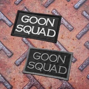 Può includere: Due toppe rettangolari nere ricamate con le parole "GOON SQUAD" in filo bianco e grigio. Le toppe sono esposte su una superficie metallica strutturata e arrugginita.