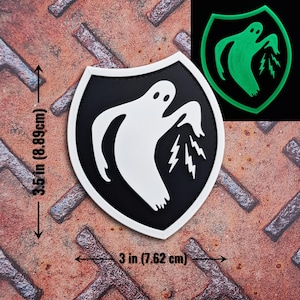 Op de afbeelding: Een zwart-wit patch met een spook- en bliksemontwerp, 8,89 cm hoog en 7,62 cm breed. Er is ook een kleinere, groen oplichtende versie afgebeeld. De patch heeft een schildvorm.