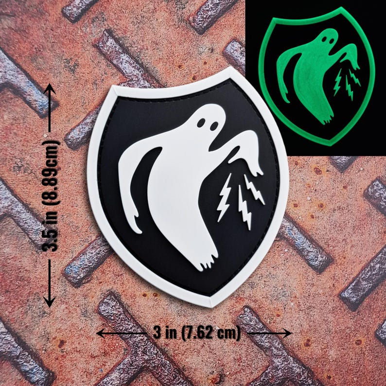 Op de afbeelding: Een zwart-wit patch met een spook- en bliksemontwerp, 8,89 cm hoog en 7,62 cm breed. Er is ook een kleinere, groen oplichtende versie afgebeeld. De patch heeft een schildvorm.