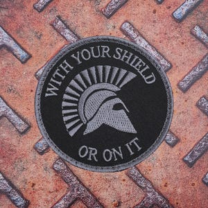 Pode incluir: Um patch redondo preto com o texto "WITH YOUR SHIELD OR ON IT" circundando um design de capacete espartano cinza. O patch tem uma borda cinza e está sobre uma superfície de metal enferrujado.