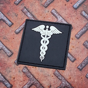 May include: A black patch with a white medical symbol, the Rod of Asclepius, featuring a snake wrapped around a staff with wings at the top.