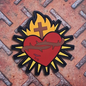 Könnte beinhalten: Ein gummiertes Abzeichen mit einem roten Herzen, das von einer braunen Dornenkrone umgeben ist, gekrönt von einem braunen Kreuz und gelben Flammen. Das Design ist von einem schwarzen Rand mit strahlenden gelben Spitzen umgeben.