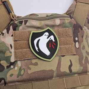 Op de afbeelding: Een camouflage tactisch vest met een patch met een wit spookontwerp op een zwart schild met een groene rand. De geest heeft rode bliksemdetails.